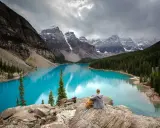 Person sitzt auf Felsen und blickt auf den türkisfarbenen Moränen-See im Banff National Park, Kanada. Majestätische Berge und Nadelwälder umgeben das Wasser.