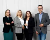 Cztery osoby w eleganckich strojach stoją przed jasną ścianą.
