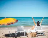Kobieta cieszy się urlopem wypoczynkowym na plaży, z rękami w górze. Obok leżaki i parasol.