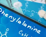 Na ekranie tabletu nazwa "Phenylalanine" i wzór chemiczny. Obok proszek fenyloalaniny w probówce i na kartce.