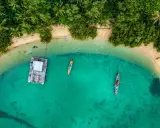 Drohne über Thailand: Ein Blick von oben auf eine schwimmende Plattform und zwei Boote im türkisfarbenen Meer, umgeben von üppiger Vegetation und Sandstrand.