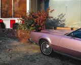 Un'auto rosa pastello, un classico stile William Eggleston, parcheggiata vicino a un muro di mattoni e un edificio con pannelli rossi e bianchi.