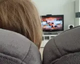 Niemowlę z blond włosami ogląda bajkę na telewizorze w tle, siedząc na kanapie.