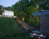 Vacker altan med decklights som lyser upp trappan och trädgården. Ett växthus syns i bakgrunden.