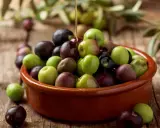 Aceite de arbequina vertiéndose sobre aceitunas verdes y moradas en un cuenco de barro. Ideal para ensaladas y aliños.