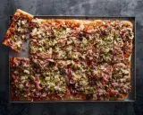 Prostokątna pizza 4 sery z pieczarkami, szynką i oliwkami, pokrojona na kwadraty, z jednym kawałkiem odłożonym na bok.