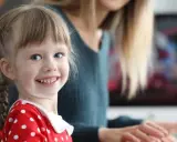 Uśmiechnięta dziewczynka w czerwonej sukience w groszki, z warkoczykami, uczy się kindersztuby od mamy.