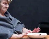 Starsza kobieta w szarym szalu odwraca się od talerza z zupą, wyraźnie odczuwając brak apetytu.
