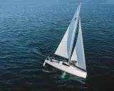 Un voilier quille relevable navigue sur une mer calme, ses voiles blanches gonflées par le vent. Deux personnes sont à bord.
