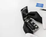 Ręce w czarnych rękawiczkach rozbierają iPhone'a, który wpadł do wody, ale działa. Widać baterię i wnętrze telefonu.