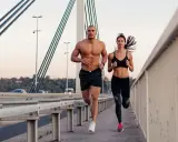 Ein Mann und eine Frau joggen auf einer Brücke. Beide haben sichtbare bauchmuskeln, ein Zeichen für ihr intensives Training.