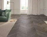 Elegante soggiorno con divano verde, lampada dorata e un tappeto beige. Il pavimento in parquet a spina di pesce, con un colore scuro e venature evidenti, completa l'arredamento.