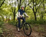 Mężczyzna w kasku jedzie rowerem trekkingowym po leśnej ścieżce. Taki rower to wszechstronny towarzysz podróży.