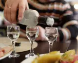 Ręka nalewa wódkę do kieliszków, przygotowując się na rekord promili. Na stole wędlina i przekąski.