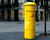 Jasnożółta skrzynka pocztowa Correos, symbol poczty Hiszpanii, stoi na brukowanym placu przed budynkami.