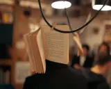 Un libro aperto, appeso per creare una scena da foto libri aesthetic. Le pagine sono piene di testo, con persone sfocate sullo sfondo.