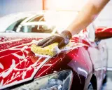 Ręka w czarnej rękawiczce myje błyszczący, czerwony samochód gąbką. Piana spływa po karoserii, tworząc efektowne smugi.