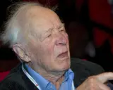 Äldre man med vitt hår, klädd i blå skjorta och mörk kavaj. Han bär ett band runt halsen med texten "Vänsterpartiet".
