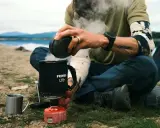 En optimus hiker njuter av en varm dryck vid en sjö. Ånga stiger från koppen medan personen sitter bredvid ett tält.