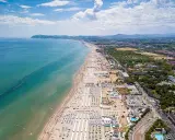 Vista aerea della Riviera Romagnola con spiaggia affollata, mare azzurro e verdeggiante entroterra.