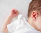 Un neonato dorme troppo, con la testolina appoggiata su un morbido lettino bianco. Si intravede un orecchio rosa e una manina.