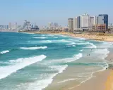 Plaża w Tel Awiwie, z falami Morza Śródziemnego i nowoczesnym miastem w tle. Ludzie kąpią się i opalają, co może sugerować, że **czy w Izraelu jest bezpiecznie** dla turystów.