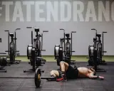 Mann liegt erschöpft auf dem Boden neben einer Hantelstange, umgeben von Trainingsgeräten. Ein Zeichen an der Wand verkündet "#TATENDRANG" – ein Symbol für den Wiedereinstieg in den **Sport nach Corona**.