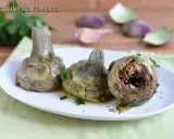 Tre carciofi alla romana cotti, serviti su un piatto bianco con prezzemolo tritato. Sullo sfondo, spicchi d'aglio e foglie di carciofo.