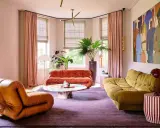 Comment amenager un salon : un fauteuil orange, un canapé rouille, un canapé moutarde, une table basse colorée, un tapis violet et une grande peinture.