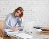 Kobieta z rudymi włosami pisze w zeszycie, obok laptop, kalkulator i książki. To jej kolejny dzień pracy nad angielskim review.