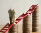 Figurki starszej pary stoją na stosach monet, połączonych czerwoną drabiną. Symbolizuje to nowe zasady przejścia na emeryturę i drogę do finansowej stabilności.