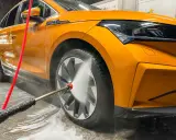 Pomarańczowe auto w trakcie mycia. Woda pod ciśnieniem spływa po karoserii i felgach, tworząc pianę.