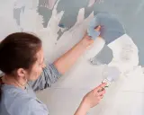 Une femme utilise une spatule pour décoller du papier peint ancien.