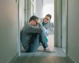 Mężczyzna w pozycji embrionalnej, z rękami na głowie, doświadcza objawów katatonii. Lekarka w białym fartuchu pochyla się nad nim, oferując wsparcie.