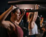 Zwei Frauen trainieren im Fitnessstudio. Sie nutzen eine Maschine, die zeigt, welche Muskelgruppen zusammen trainieren.