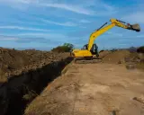 Excavadora amarilla trabajando en la creación de varios tipos de zanjas, con montones de tierra y tuberías grandes en el fondo.