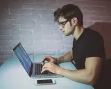 Młody mężczyzna w okularach pisze kod na laptopie. To jego pierwszy krok, by dowiedzieć się, jak zostać informatykiem.