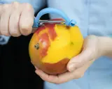 Hände schälen eine reife Mango mit einem blauen Sparschäler. Das Fruchtfleisch leuchtet gelb, während die Schale rot und gelb ist.