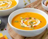 Velouté de courge butternut orange servi dans un bol blanc, garni de crème, de graines de courge et de poivre rose.