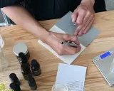 Person skriver recept för ekologiska doftpinnar. Ingredienser och verktyg ligger på ett träbord.