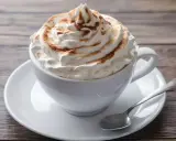 Ein cremiger Kapuziner Kaffee, üppig garniert mit Schlagsahne und Zimt, wartet auf einem Holztisch.