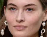Primer plano de un rostro femenino con pecas naturales salpicando su piel. Lleva pendientes de perlas colgantes.
