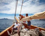 Trois amis profitent du soleil sur un voilier, rêvant d'un tour du monde. L'eau bleue et les falaises en arrière-plan ajoutent à l'aventure.