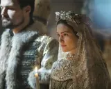 Alicia Agneson i en brudklänning med krona och slöja, bredvid en man i päls och guldbrokad. Ljus från stearinljus.