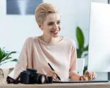 Uśmiechnięta kobieta z krótkimi blond włosami pracuje przy komputerze, obok aparat fotograficzny. Sprawdza najnowsze oferty pracy w Pile.