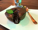 Un délicieux mi cuit chocolat coulant, garni d'une feuille de menthe, sur une assiette blanche. Une cuillère dorée est à côté.