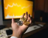 Dłoń trzyma trzy monety kryptowalut: Bitcoin, Ethereum i inną. W tle wykres na monitorze, sugerujący handel na kantory kryptowalut.