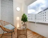 Przytulny balkon z drewna, z wiklinowym fotelem, stolikiem i rośliną. W tle białe budynki.