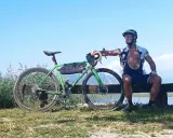 Ciclista riposa accanto alla sua bici verde, pensando alle tabelle per dimagrire in bicicletta. Cielo azzurro e lago sullo sfondo.