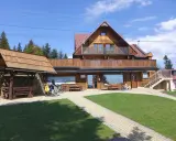 Drewniana bacówka na obidzy z balkonem i tarasem, otoczona zielenią i ścieżkami. Obok wózek inwalidzki z żółtą torbą.